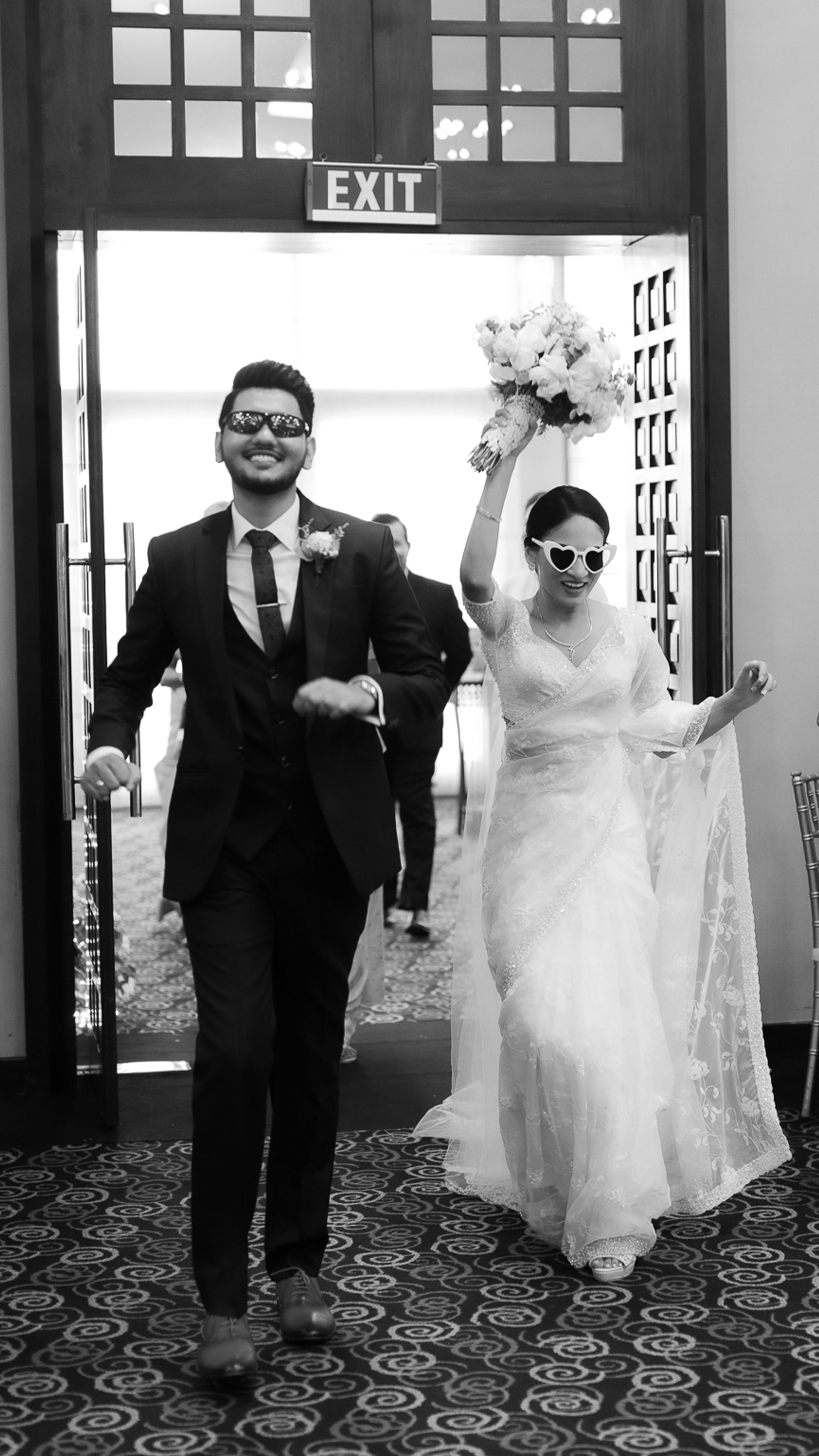 A joyful Sri Lankan bride and groom wearing sunglasses exit a venue, showcasing their unique style.