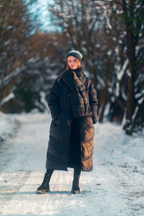 Woman in Puffer Coat and Scarf Standing on Snowy Forest Road