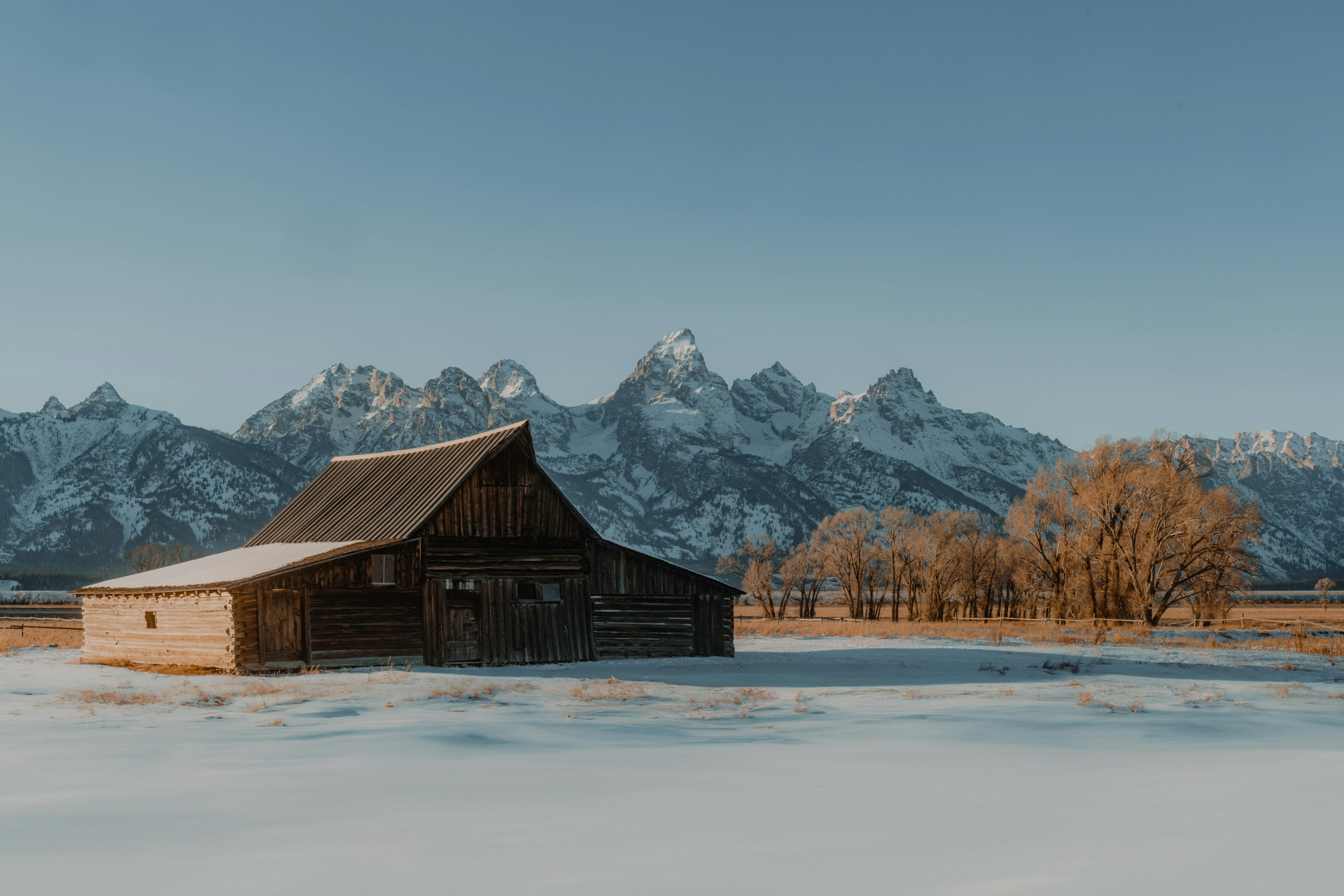 A scenic view of a rustic wooden barn against a snowy mountain range under clear blue skies.