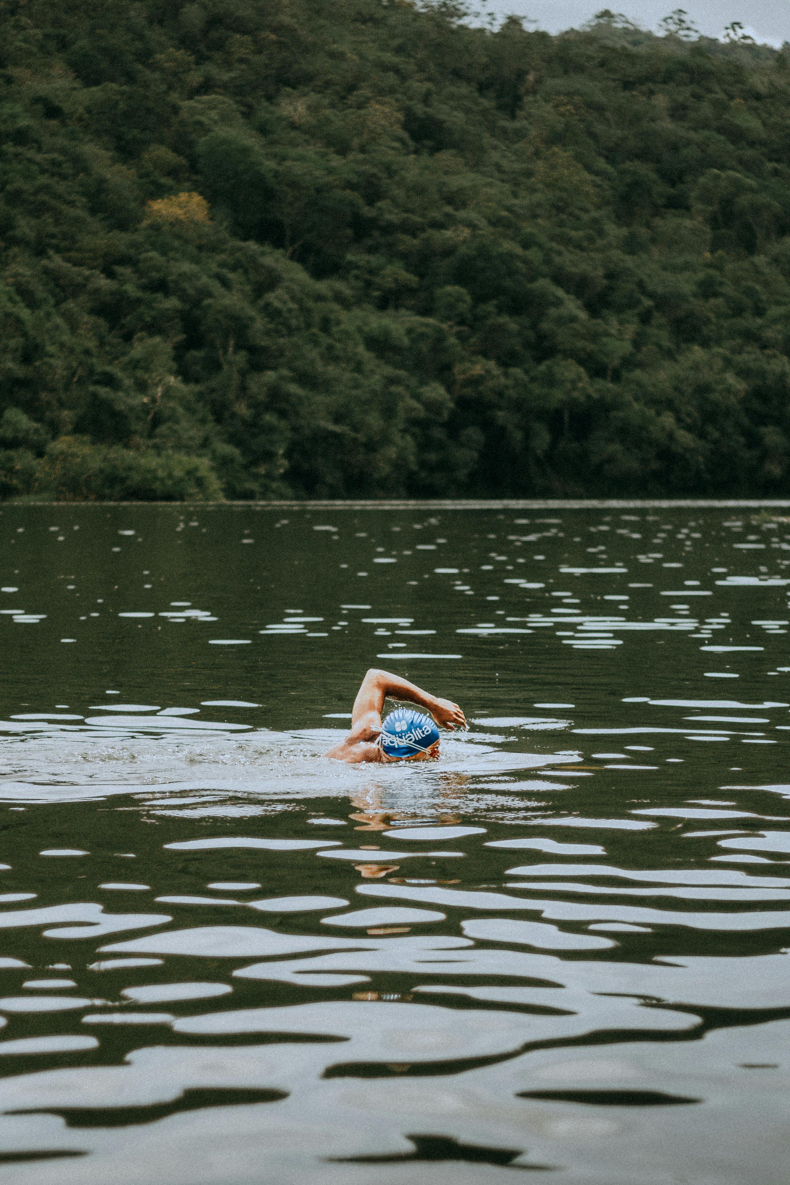 An adult swimmer wearing a blue swim cap trains in a serene forest lake.