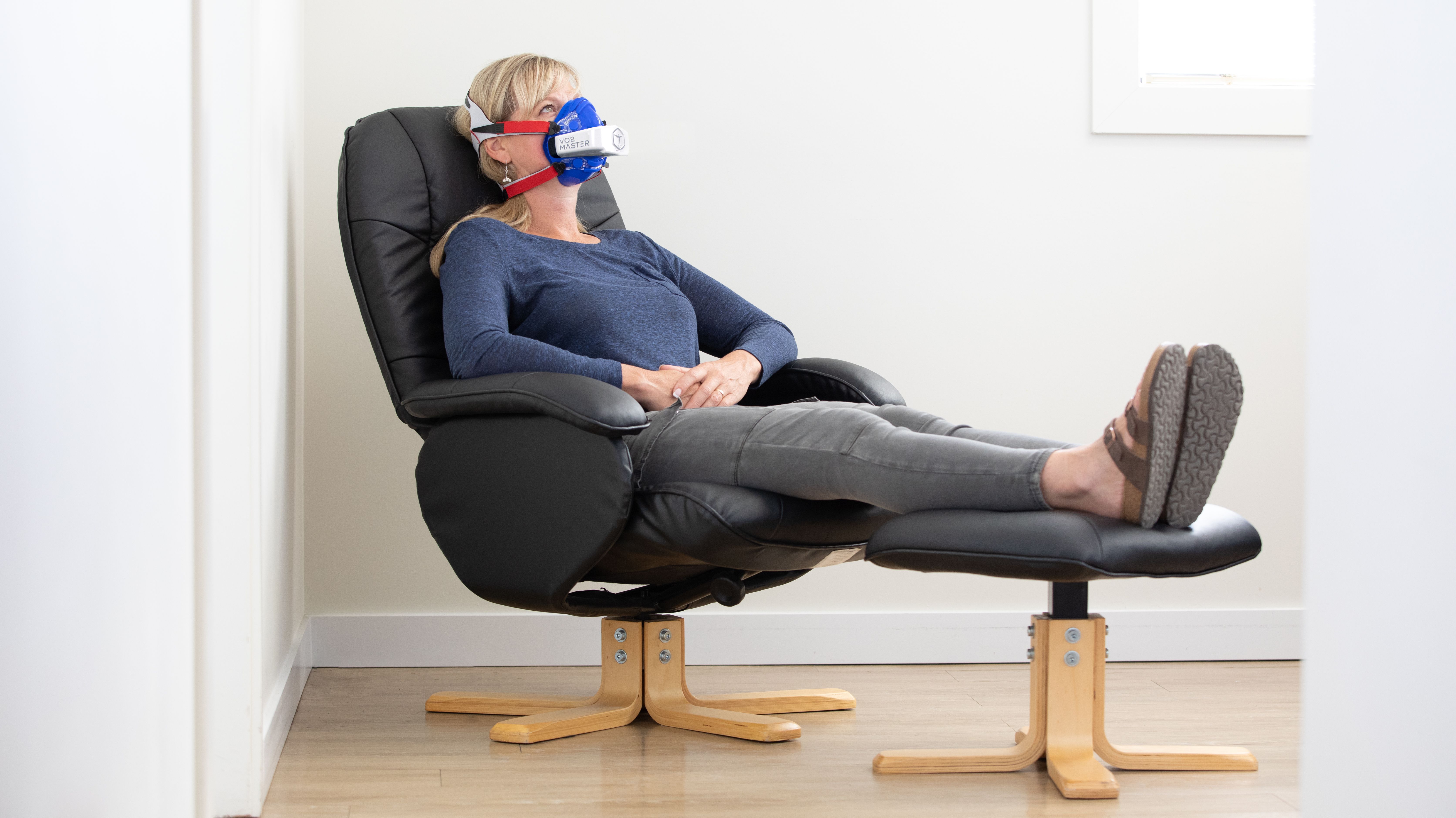 A woman comfortably reclines in a modern armchair, using a protective face mask indoors.