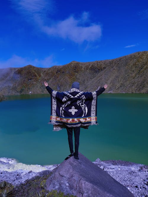 Person in Poncho Standing by Lake with Raised Hands