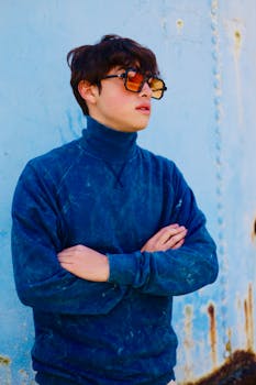 Portrait of a young man standing against a textured blue wall wearing sunglasses and a navy blue pullover.