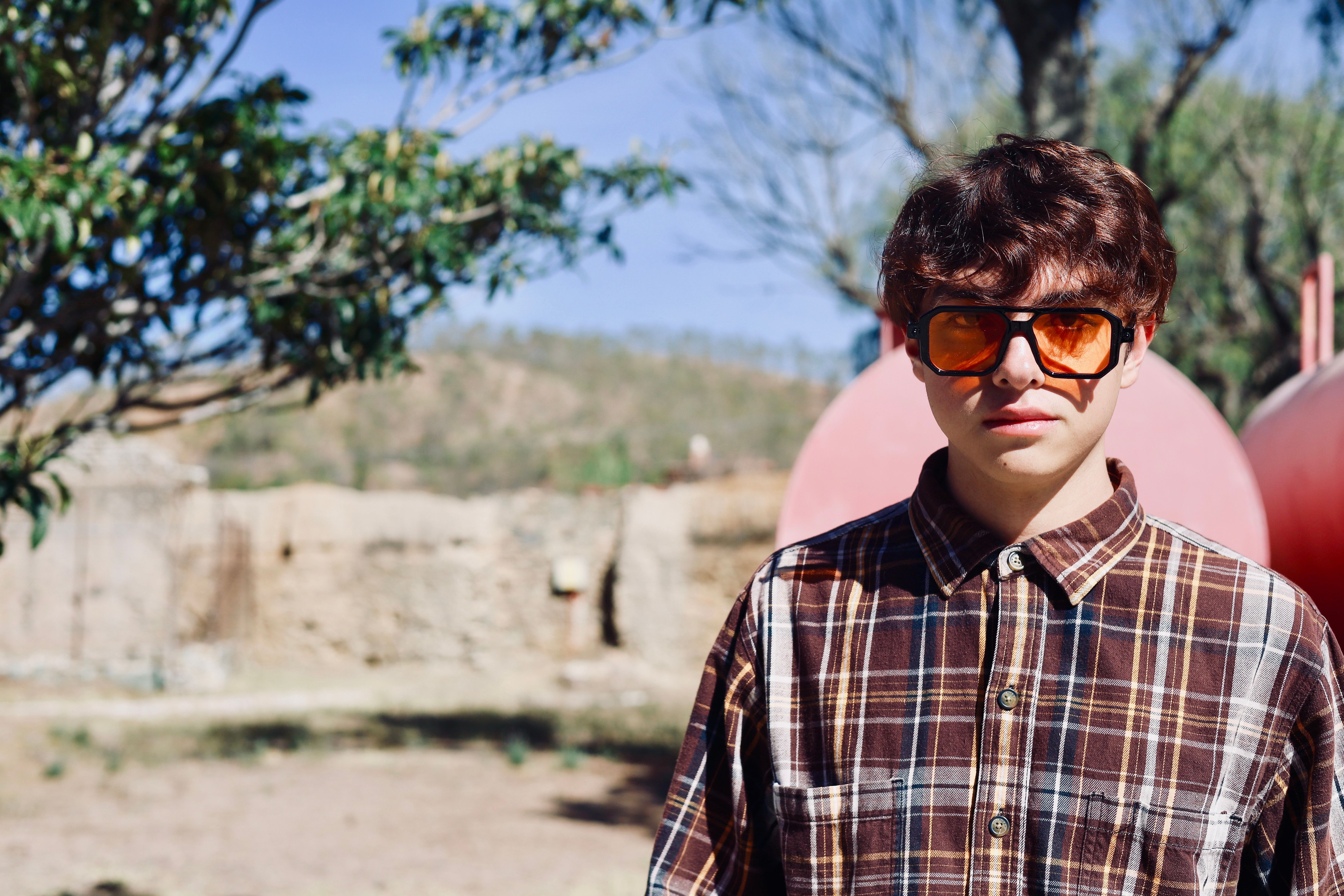 Teenager with curly hair in plaid shirt and orange sunglasses, outdoors on a sunny day.