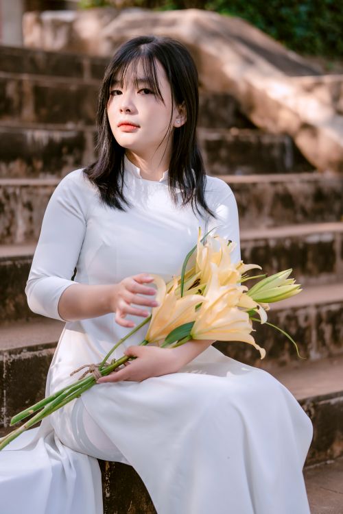 Free A woman in a white dress elegantly holds a bouquet of lilies while sitting on stairs outdoors. Stock Photo