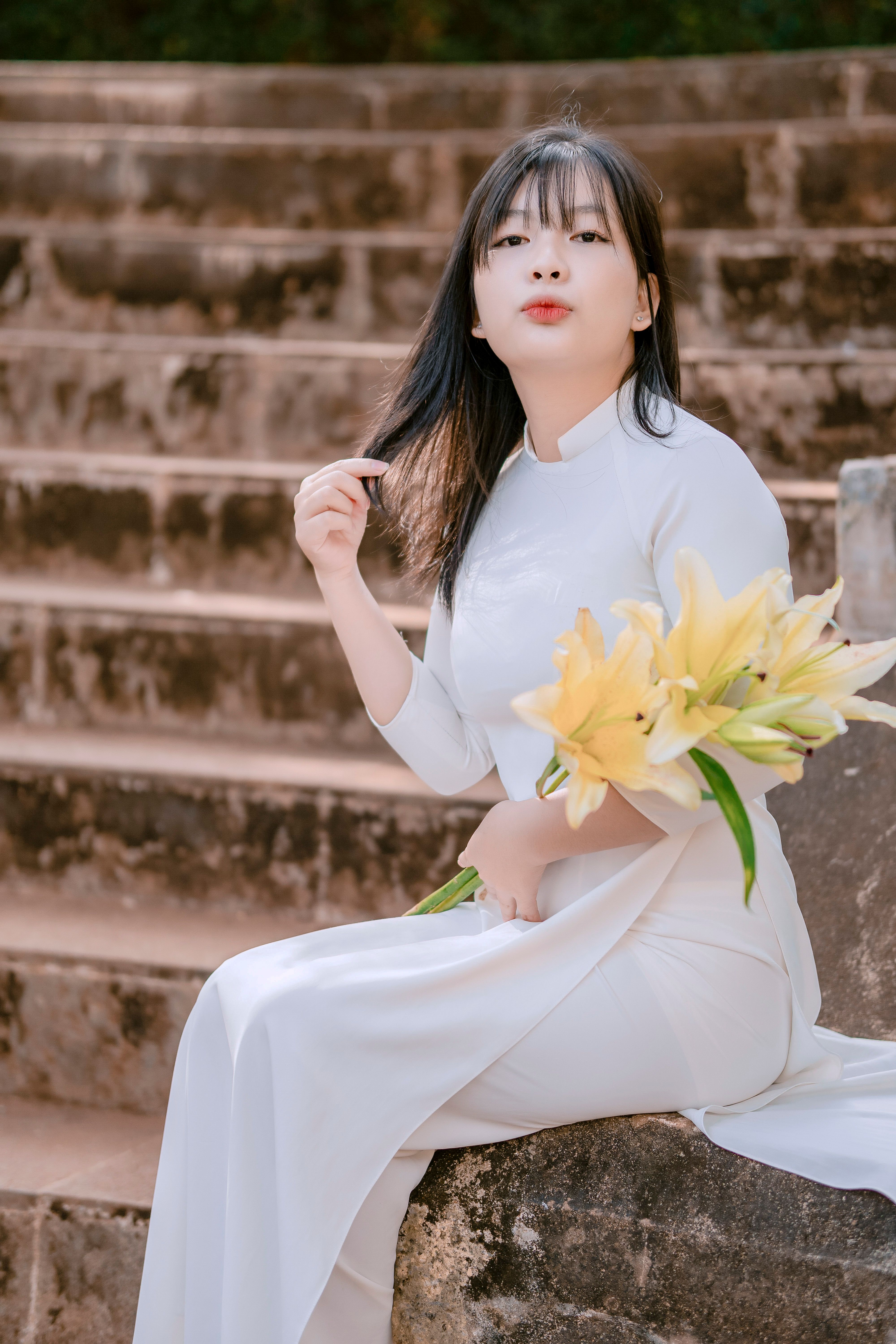An elegant Asian woman sits gracefully on steps holding a bouquet of flowers, showcasing traditional clothing.