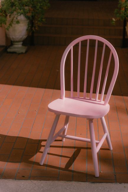 A solitary pink wooden chair casts a shadow on a sunlit patio with brick flooring.