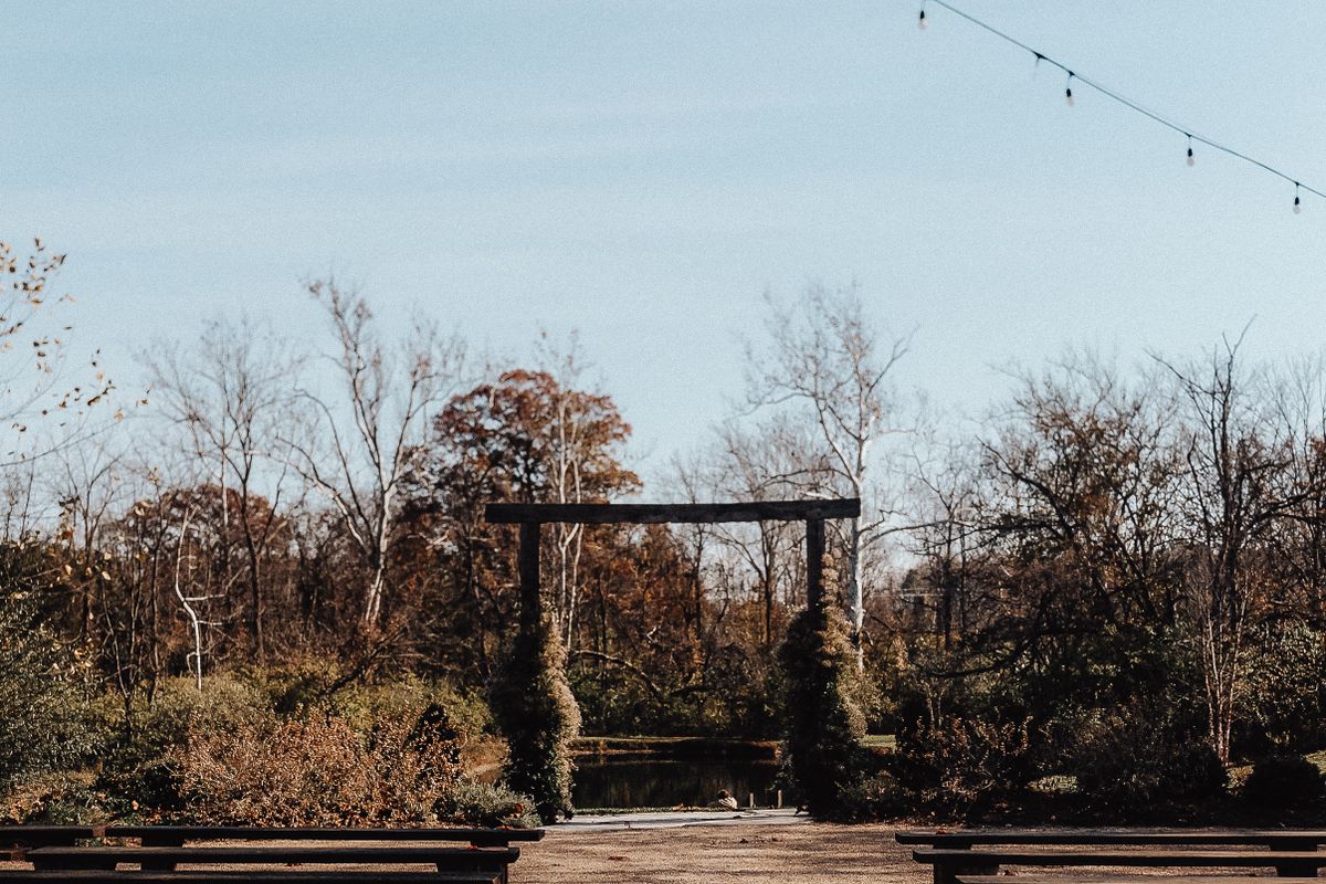 Outdoor autumn wedding ceremony venue with wooden benches, rustic arch, and fall trees in the background