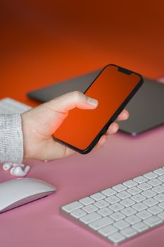 Close-up of a hand holding a smartphone over a pink desk setup with a laptop and keyboard.