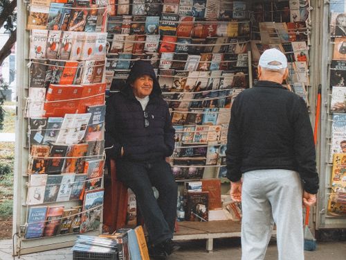 Free A bustling urban newsstand with two men conversing, surrounded by magazines and newspapers. Stock Photo