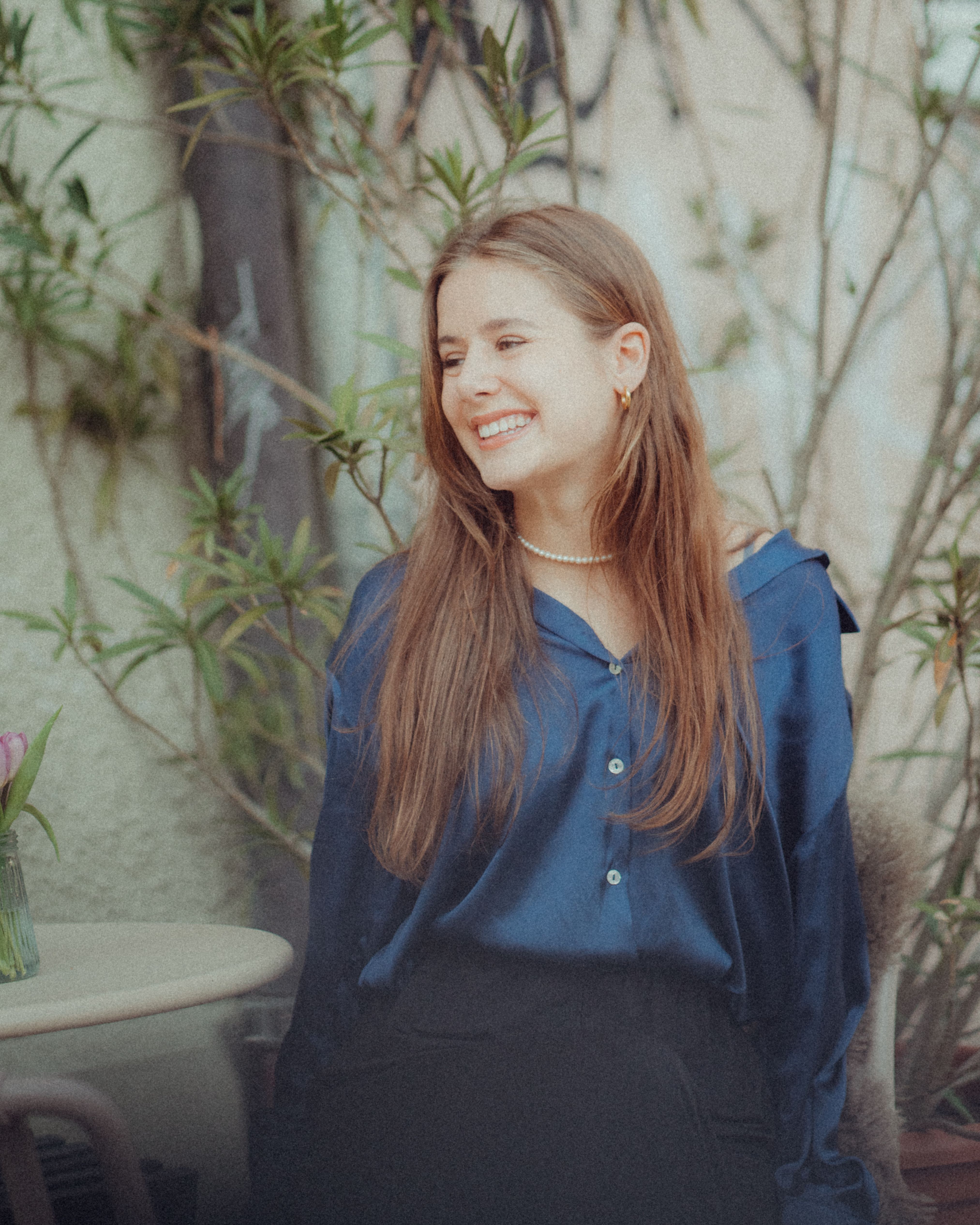 A young woman in a blue blouse happily smiling while sitting outdoors surrounded by greenery.