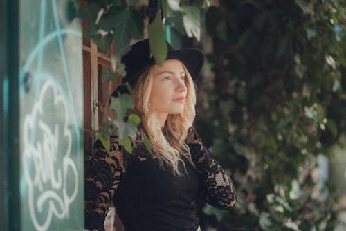 A woman in a black hat and black dress leaning against a wall