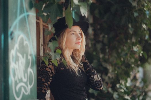 A young woman in a black hat with lace sleeves poses confidently outdoors amidst greenery.