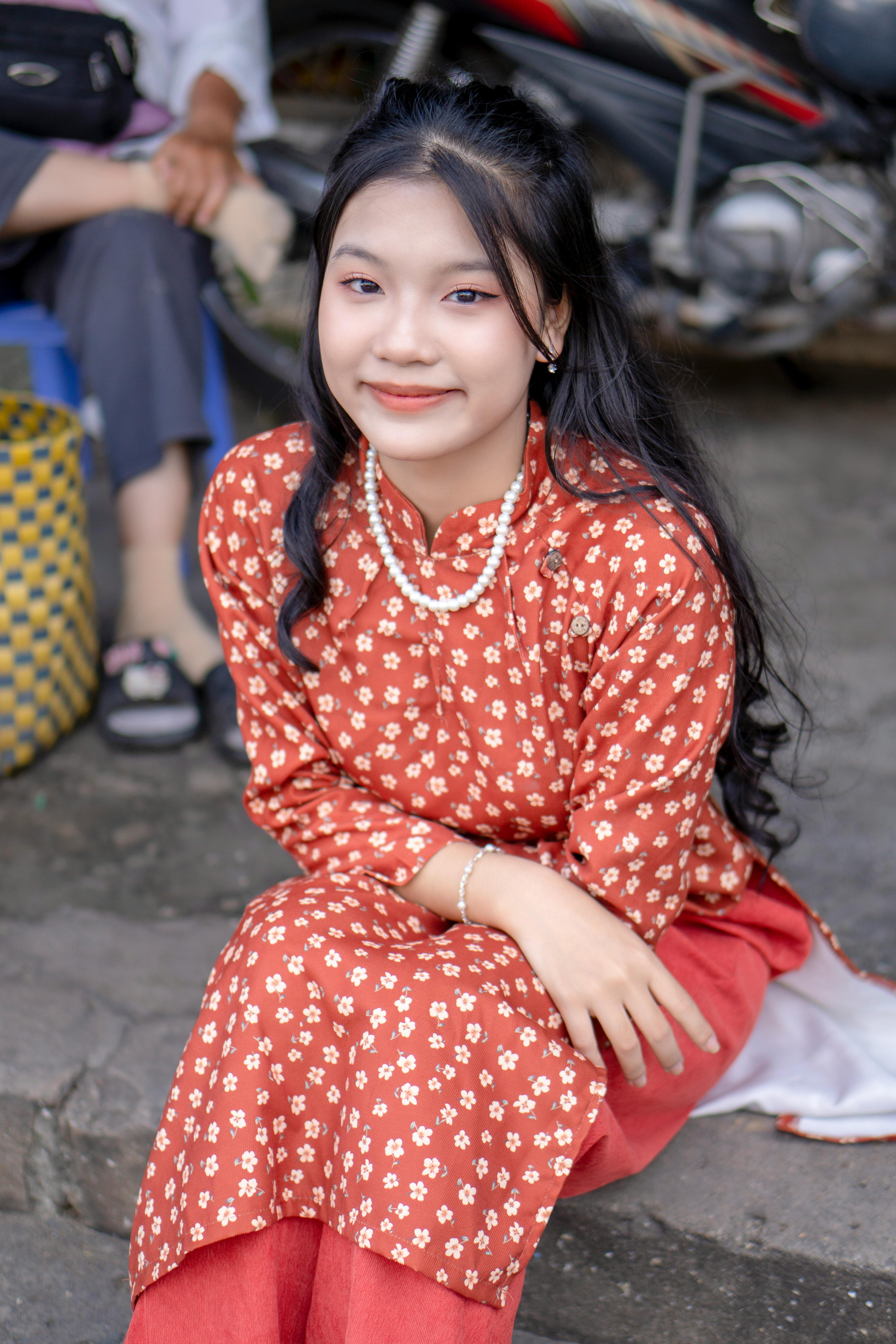 Smiling young woman in a floral tunic sitting outdoors on a street.