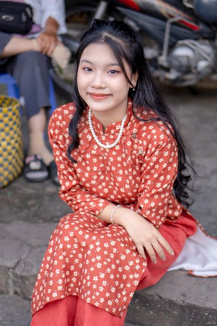 Smiling young woman in a floral tunic sitting outdoors on a street.