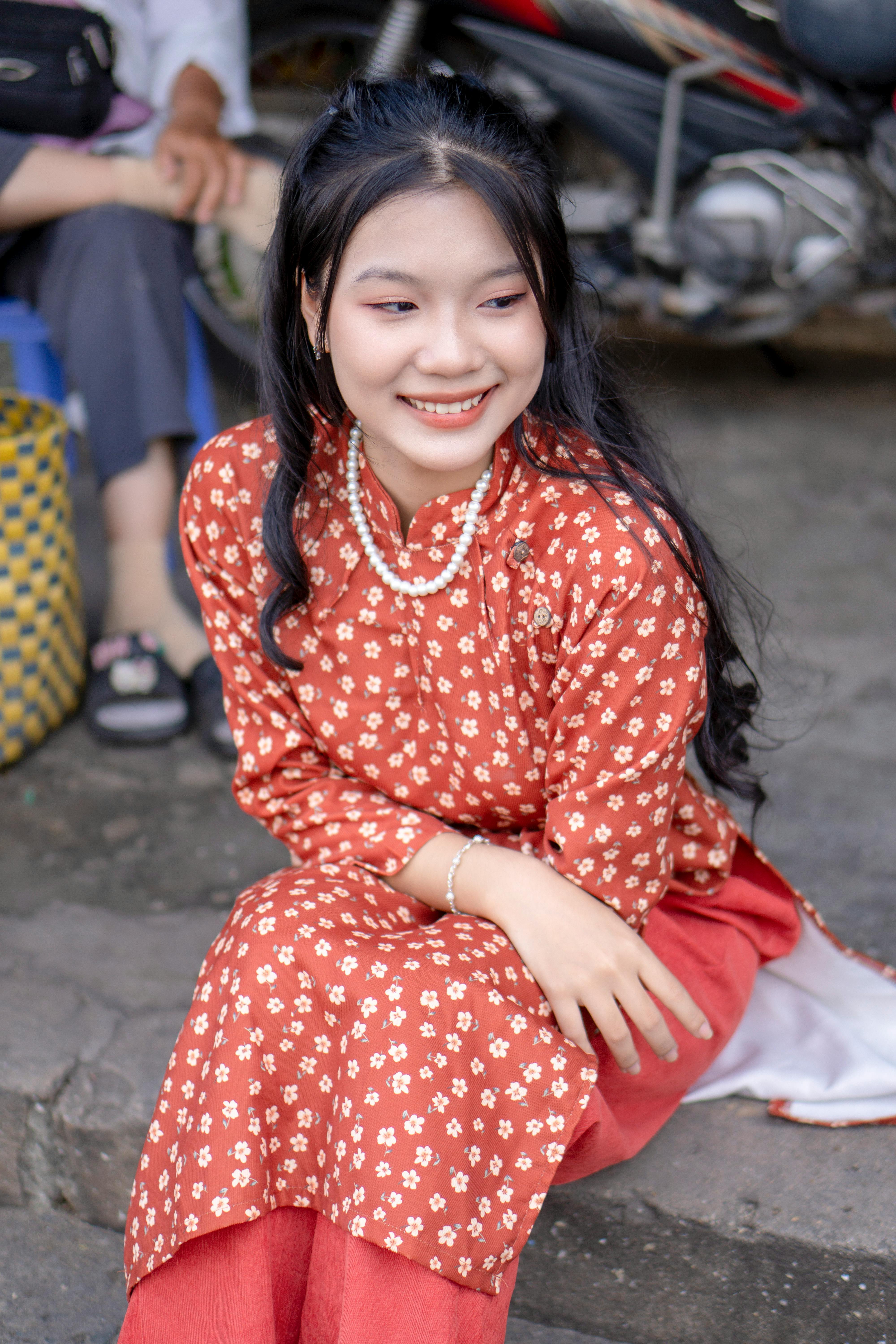 Smiling Asian woman in traditional red floral clothing sitting outdoors with a joyful expression.