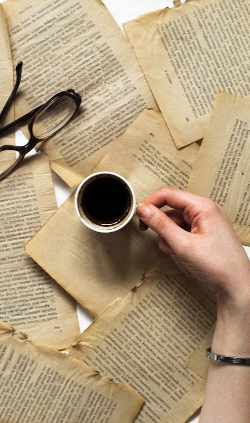 A person's hand holding a cup of coffee over a pile of books