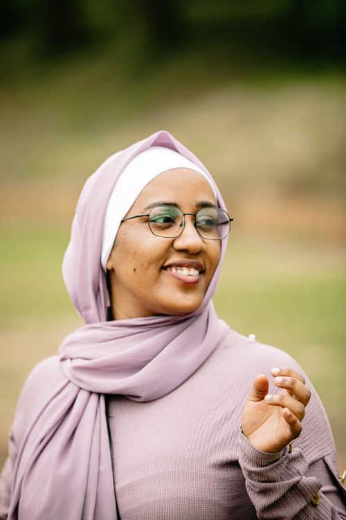 Free A Muslim woman in a hijab, wearing eyeglasses, smiles warmly in an outdoor setting, exuding positivity and style. Stock Photo