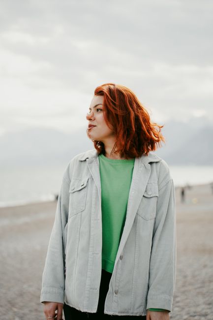 Young woman with red hair in a denim jacket at a serene beach setting, exuding a fashionable vibe.