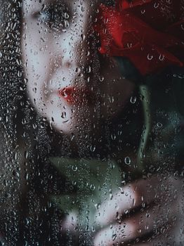 Woman holding a rose behind a rain-splattered window, creating a moody romantic scene.