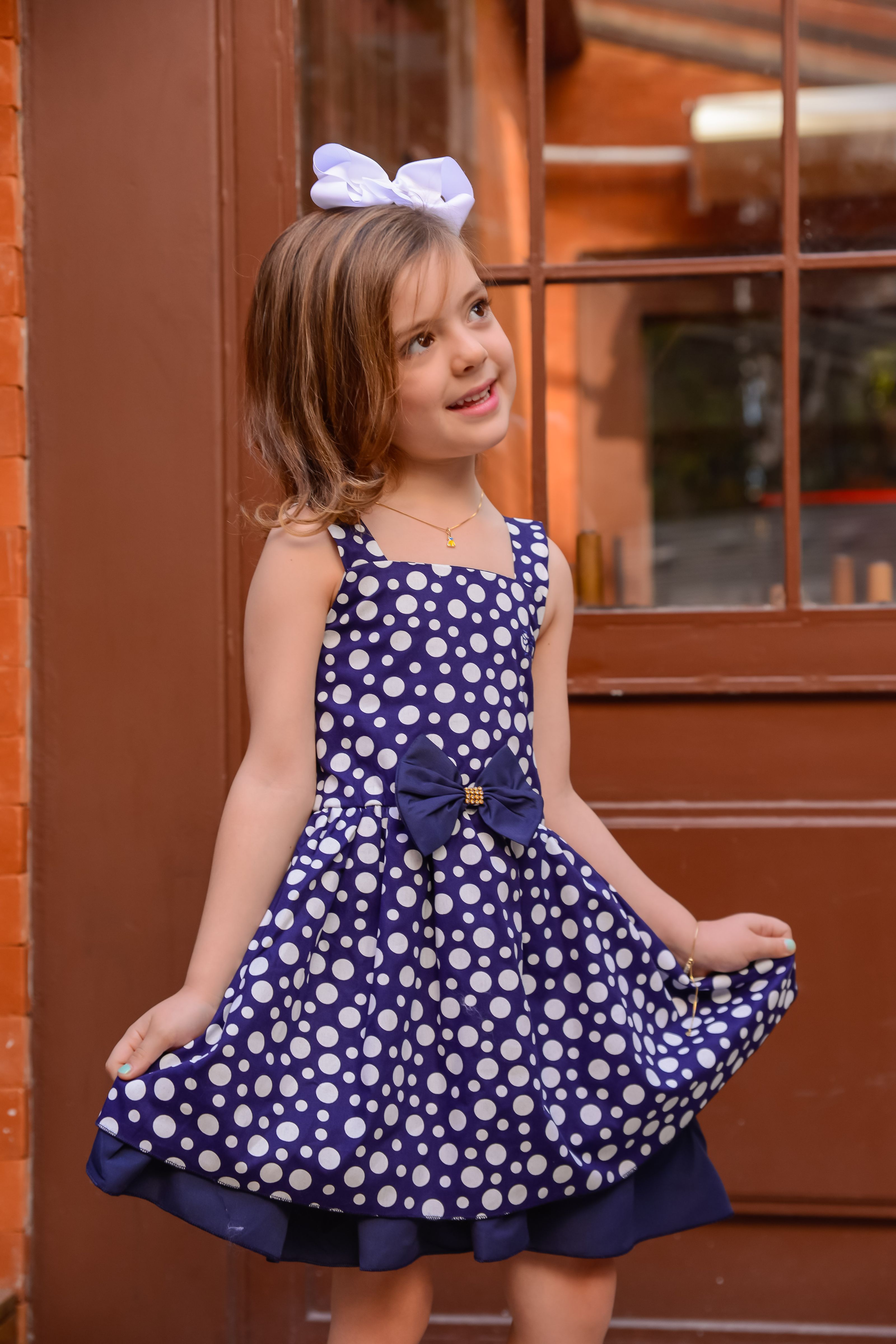 Menina encantadora sorrindo e posando em um vestido azul com bolinhas brancas, laço e fita de cabelo branca.