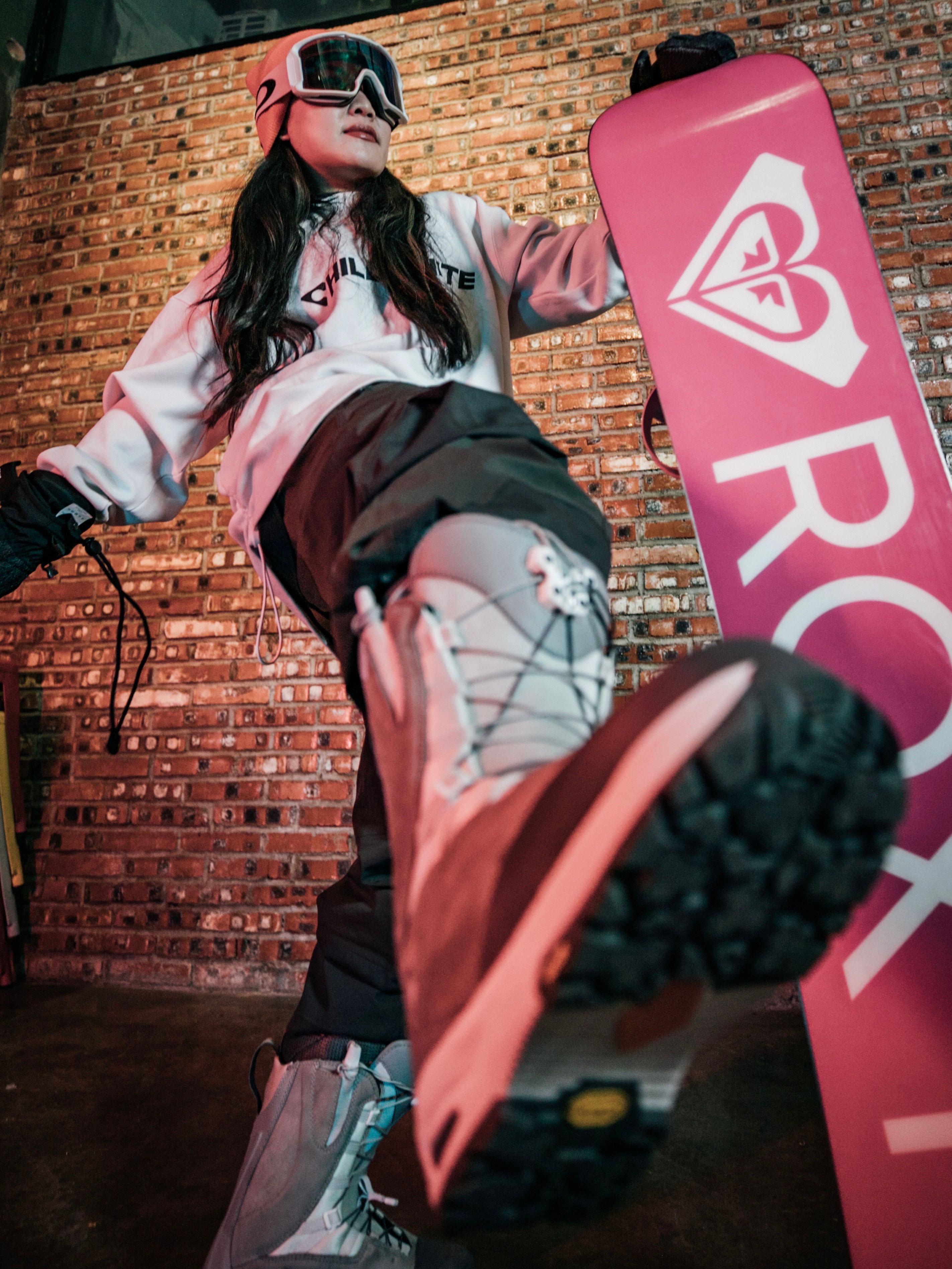 A woman snowboarder indoors with goggles and a snowboard, striking a dynamic pose.