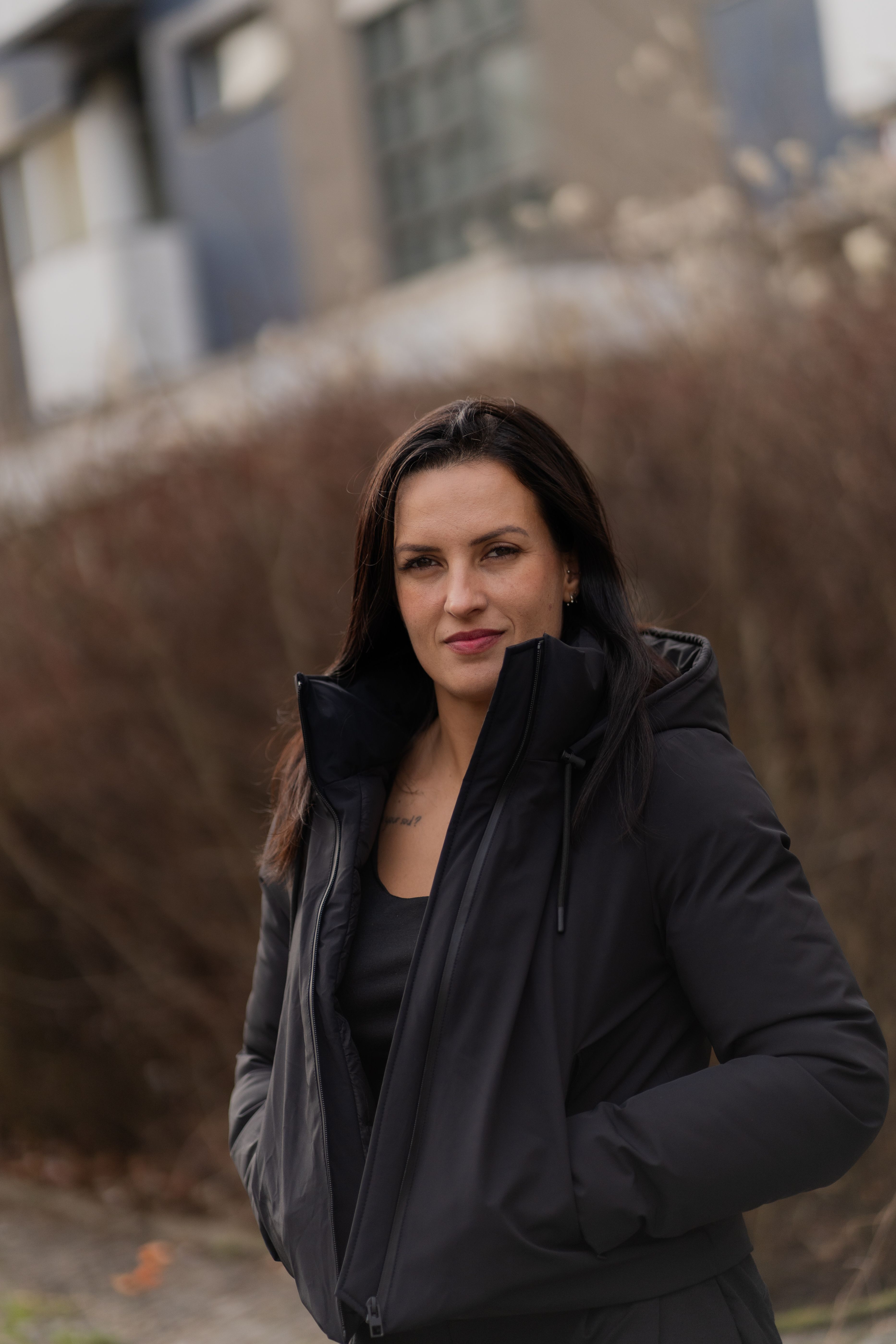 A woman stands outdoors wearing a stylish black jacket, hands in pockets.