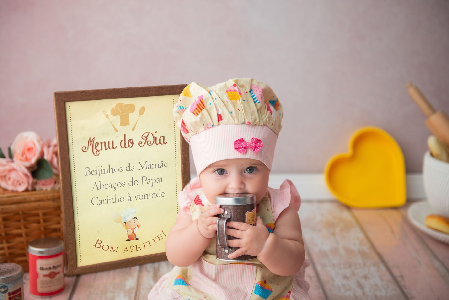 Cute baby girl dressed as a chef holding a cup, with a Portuguese menu sign in the background.
