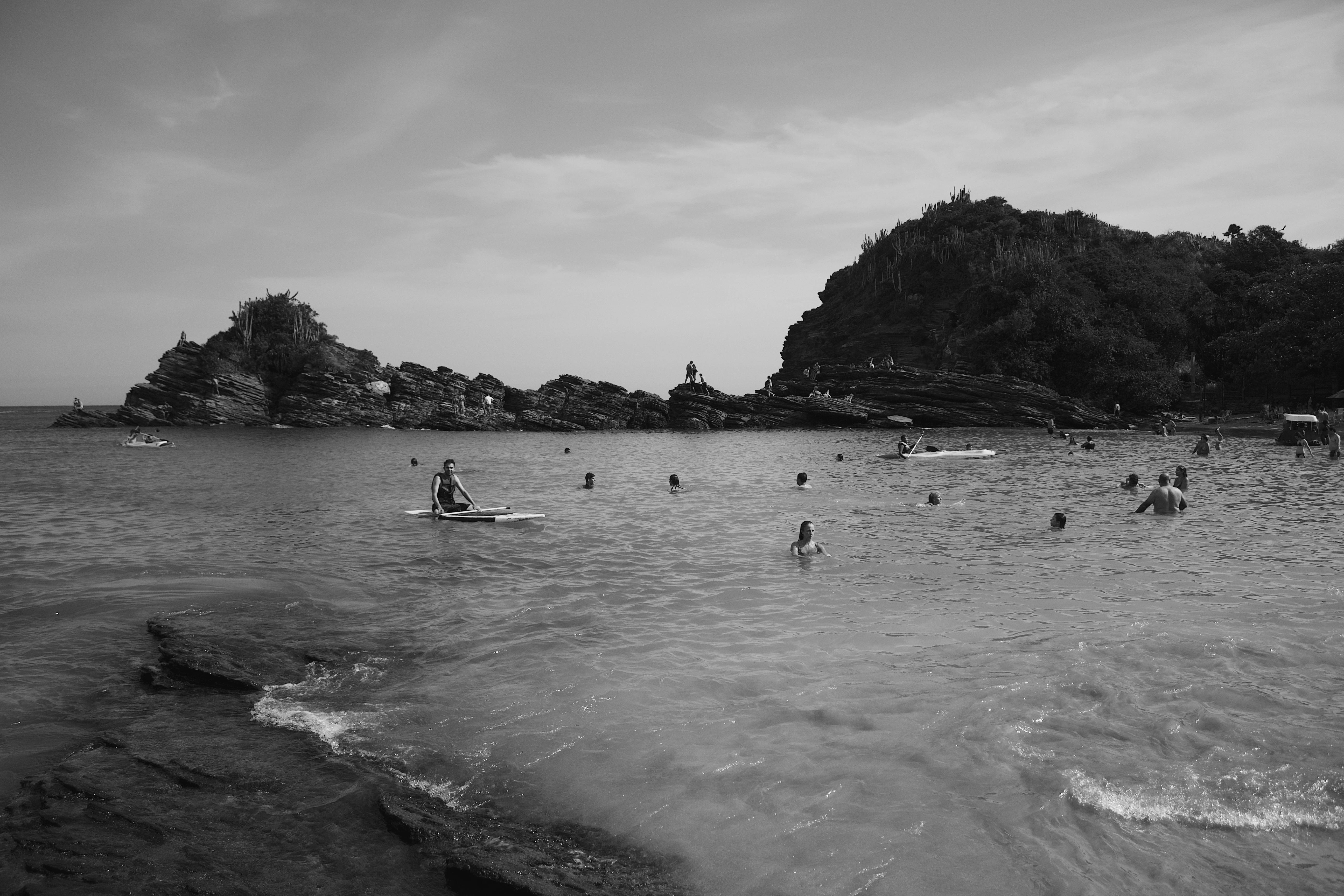 Black and white photo of a lively beach scene at Armação dos Búzios, Brazil, featuring rocky shore and swimmers.