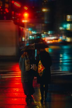 Two women with umbrellas walking through colorful, rainy New York streets at night.