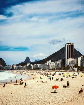 Vibrant summer scene at Copacabana Beach with crowds, umbrellas, and city skyline.