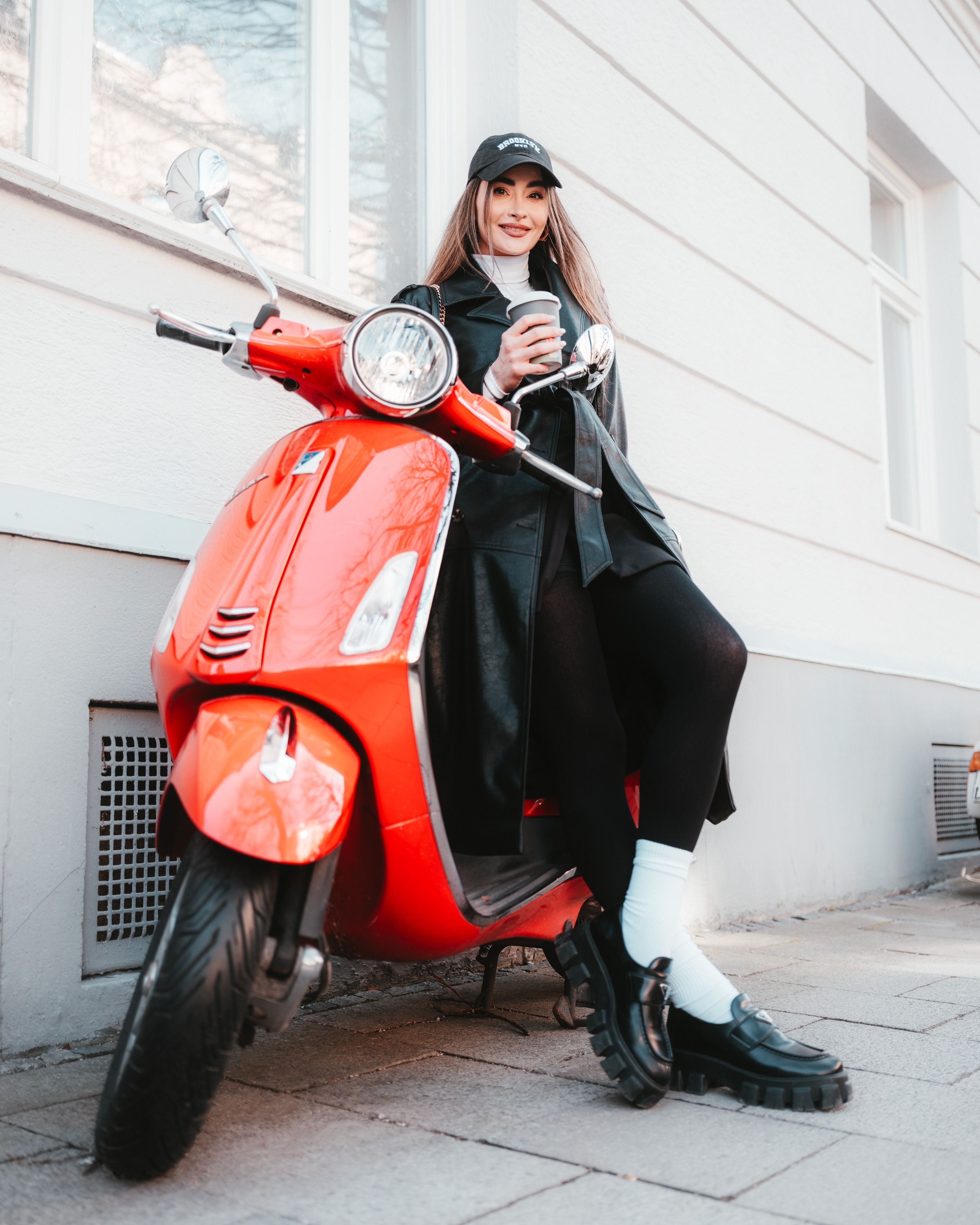 Fashionable woman in black coat and hat sits on a red scooter outside a city building holding a drink.