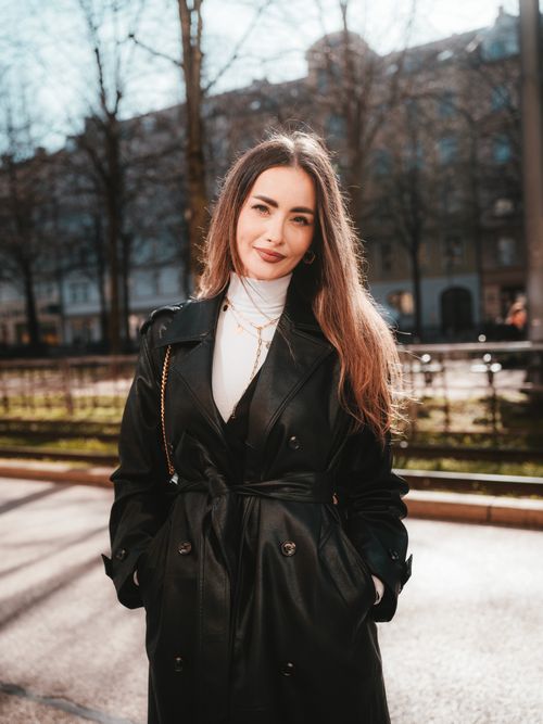 Free Woman in black coat poses on a sunlit city street, showcasing urban fashion and confidence. Stock Photo