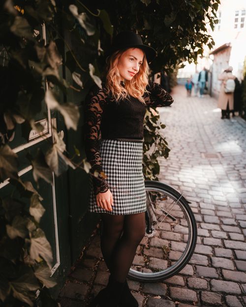 Free Stylish woman with blonde hair poses against a leafy wall in a sunlit cobblestone alley. Stock Photo