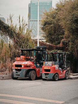 Two forklifts parked on a city street, surrounded by greenery and urban buildings.