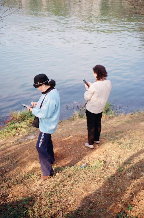 Two people standing near a body of water with cell phones