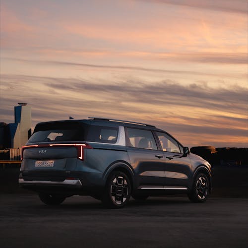 The right rear view of the Kia Carnival Hybrid parked with a building in the background under the sunset.