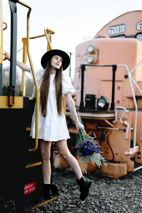Woman in Hat and White Dress Standing on Locomotive Stairs