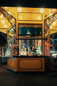 Street vendor in a bright kiosk selling food at night in a bustling city street with decorative lighting.