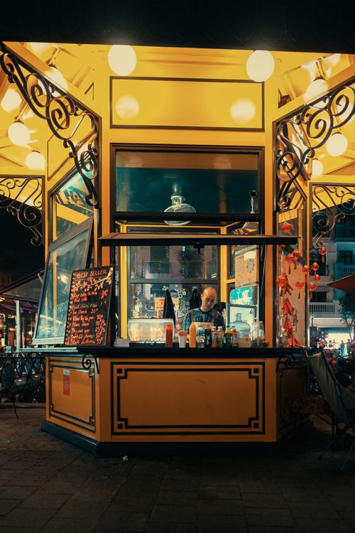 Free Street vendor in a bright kiosk selling food at night in a bustling city street with decorative lighting. Stock Photo