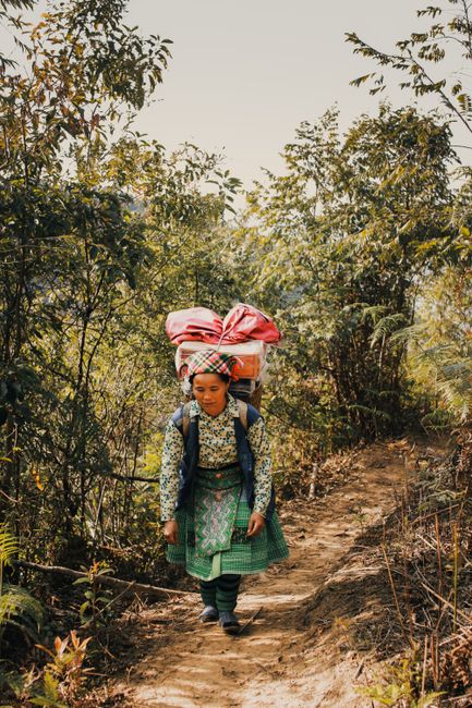 Elderly woman in traditional clothing walking a rural path with a large backpack, surrounded by trees.
