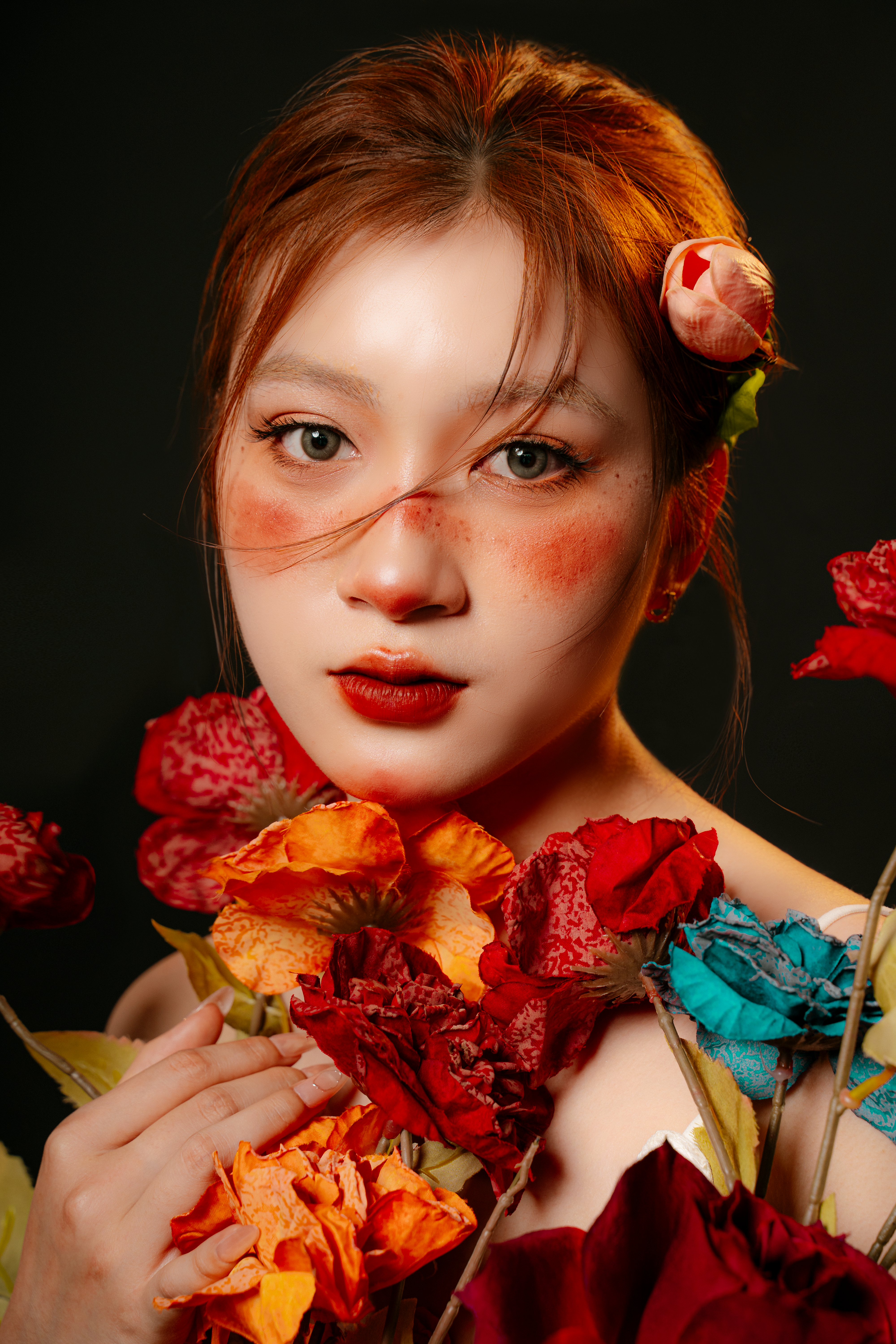 Close-up portrait of a woman surrounded by vibrant flowers in a studio setting, showcasing makeup artistry.
