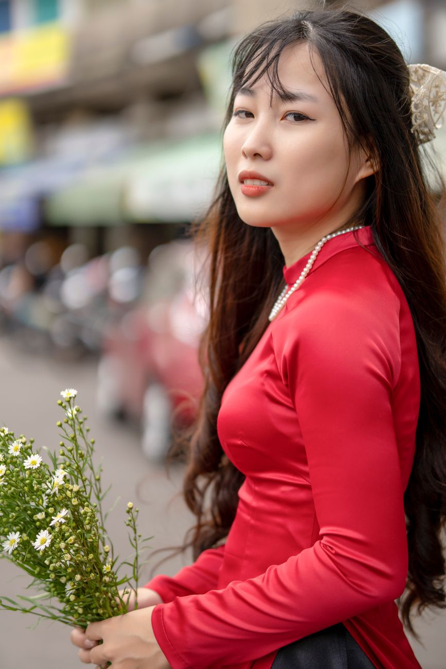 A woman in a vibrant red traditional gown poses gracefully with flowers on a bustling city street.