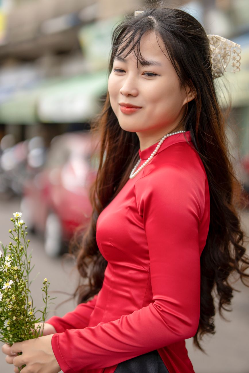 Beautiful young woman in a red dress smiling while holding flowers outdoors.