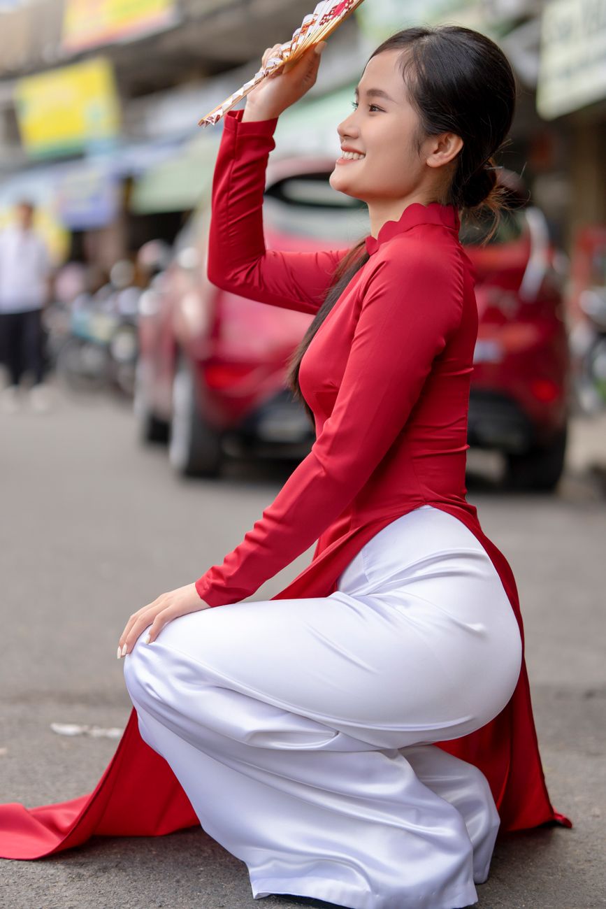 Young woman in red ao dai smiles while posing on a vibrant city street.