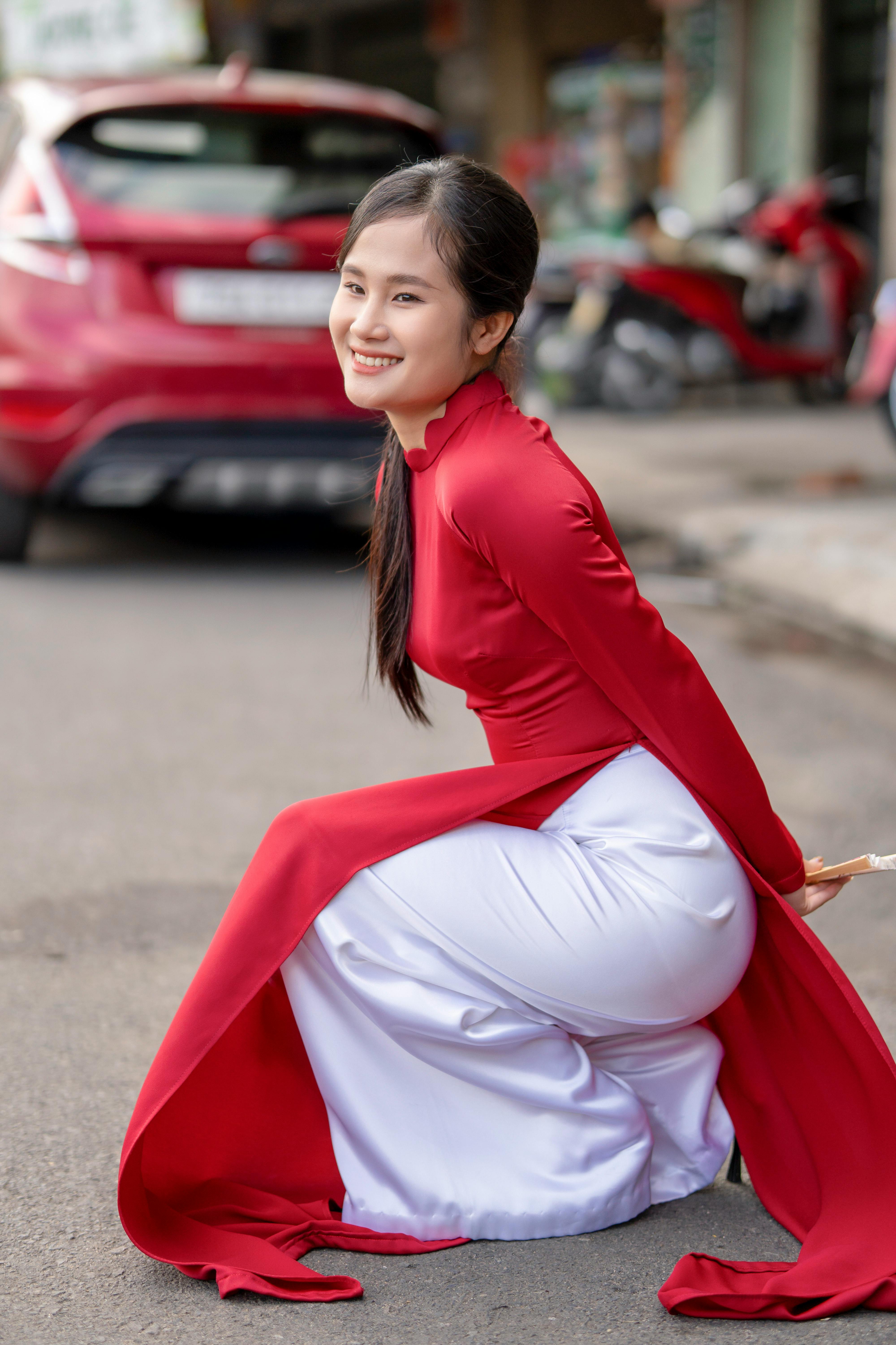 Young woman in red ao dai sitting on a city street with a vibrant smile, embodying cultural elegance.