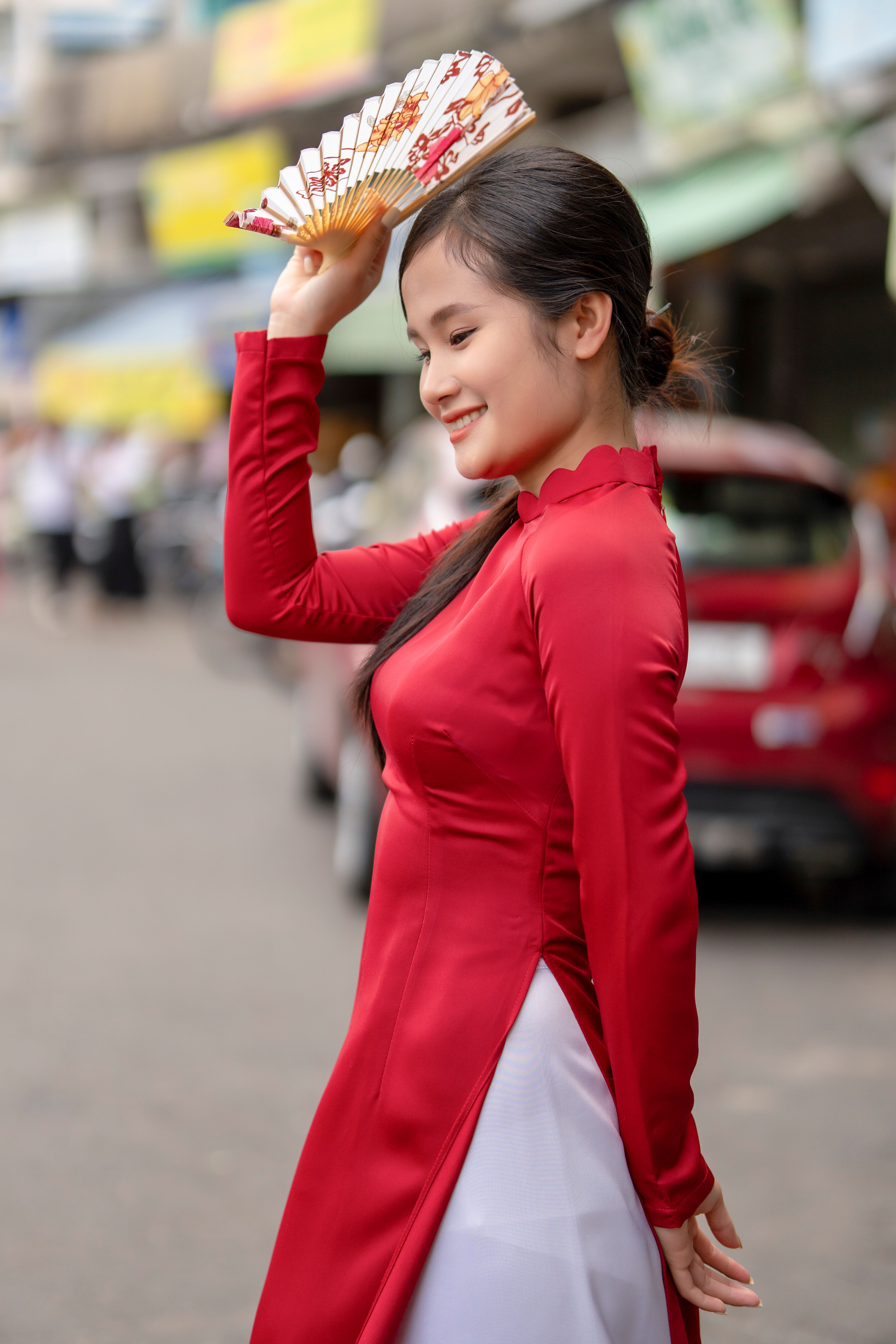 Smiling woman in red dress poses with a hand fan on city street.