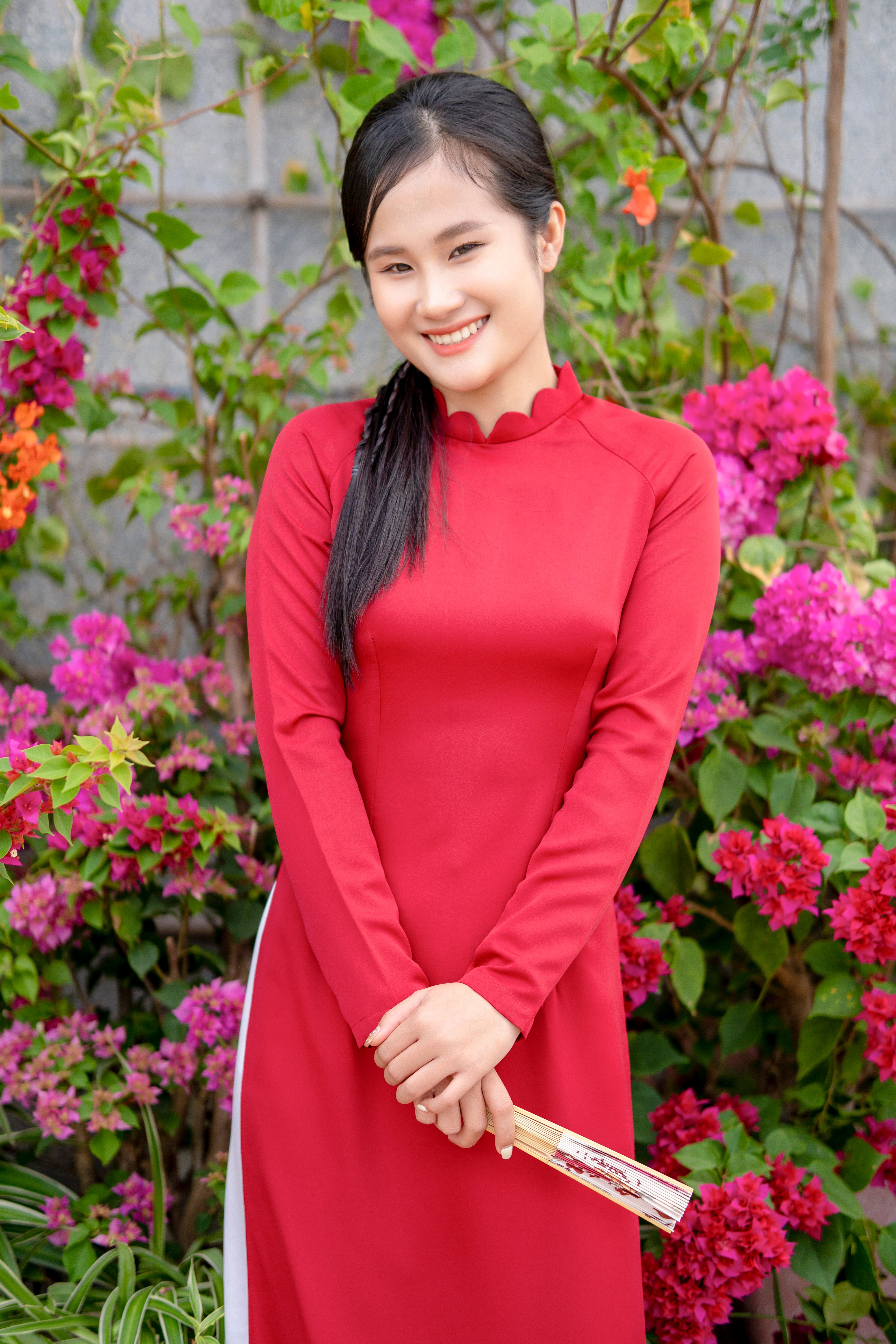A cheerful woman in a red dress smiles amidst vibrant pink flowers in a garden setting.