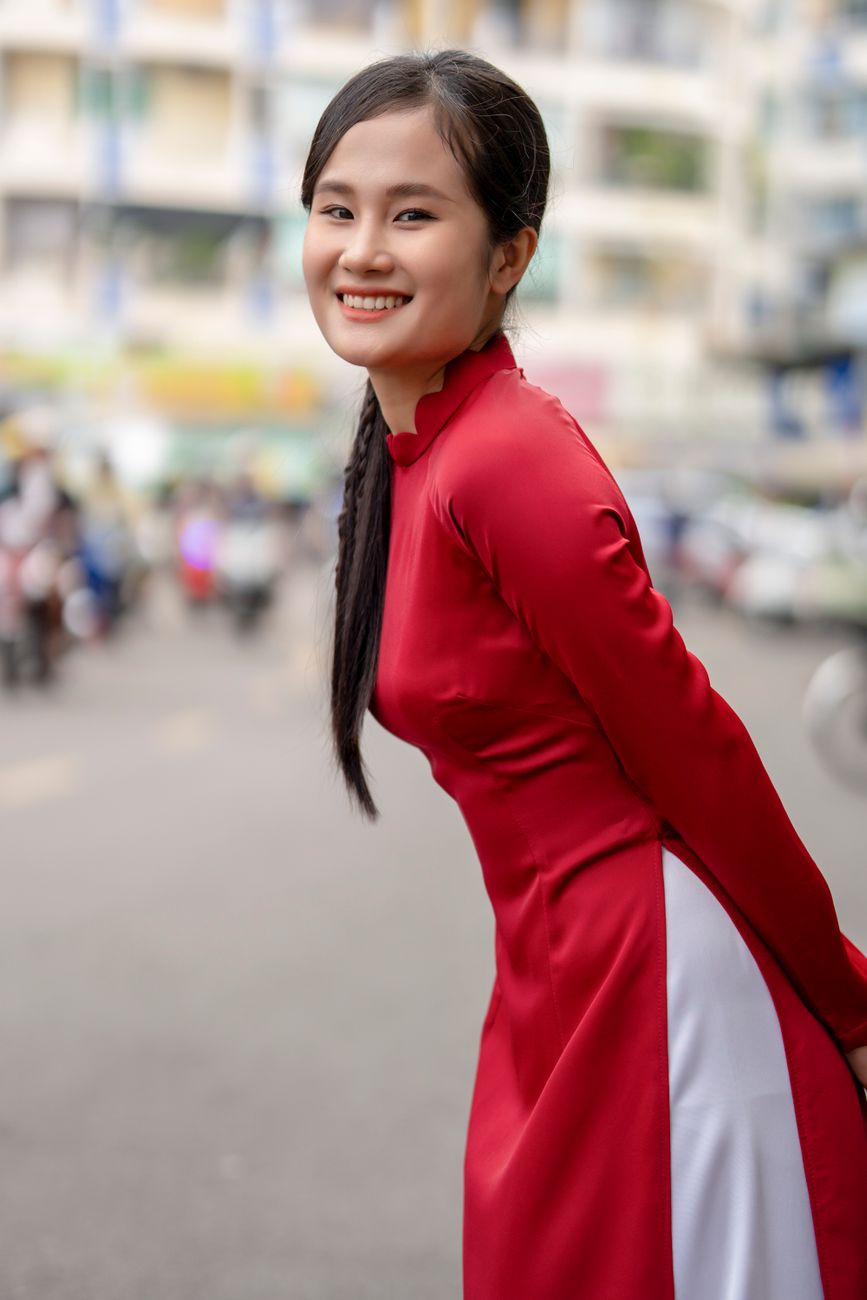 Smiling woman wearing a red traditional gown, posing on a bustling city street.