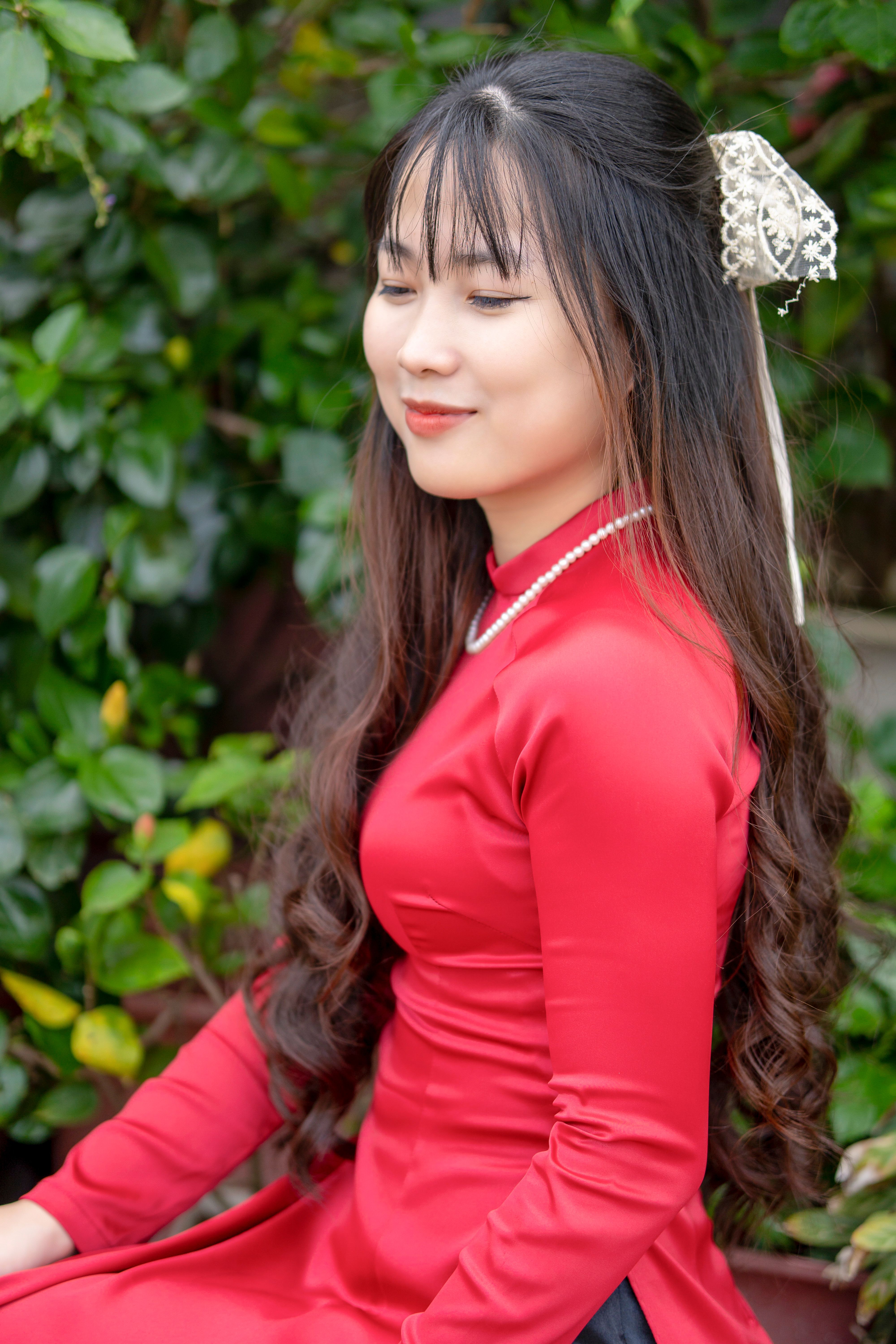A beautiful woman with long black hair smiles elegantly in a red dress against lush greenery.
